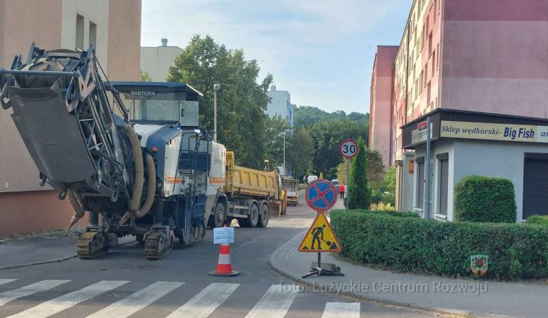 maszyna drogowa stoi po lewej stronie, po prawej widać drogę i blok mieszkalny